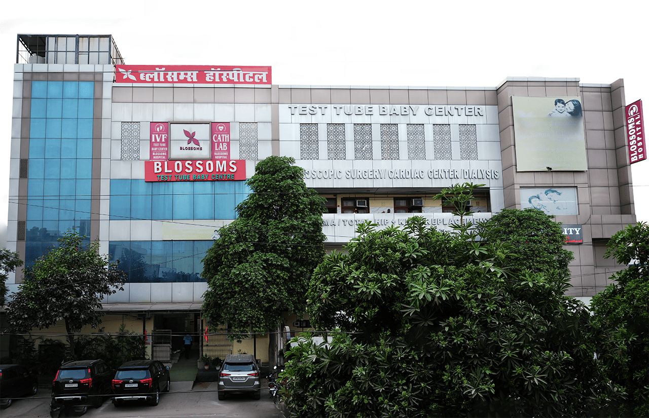 Exterior view of Blossoms Hospital, showcasing its modern building with signs for IVF, Test Tube Baby Center, and other specialized medical services, surrounded by lush greenery and parked cars.